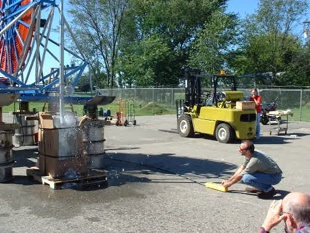 On the left is the drill portion of the contraption, spewing out water onto asphalt. there is a person in a blue shirt crouching on the ground.
