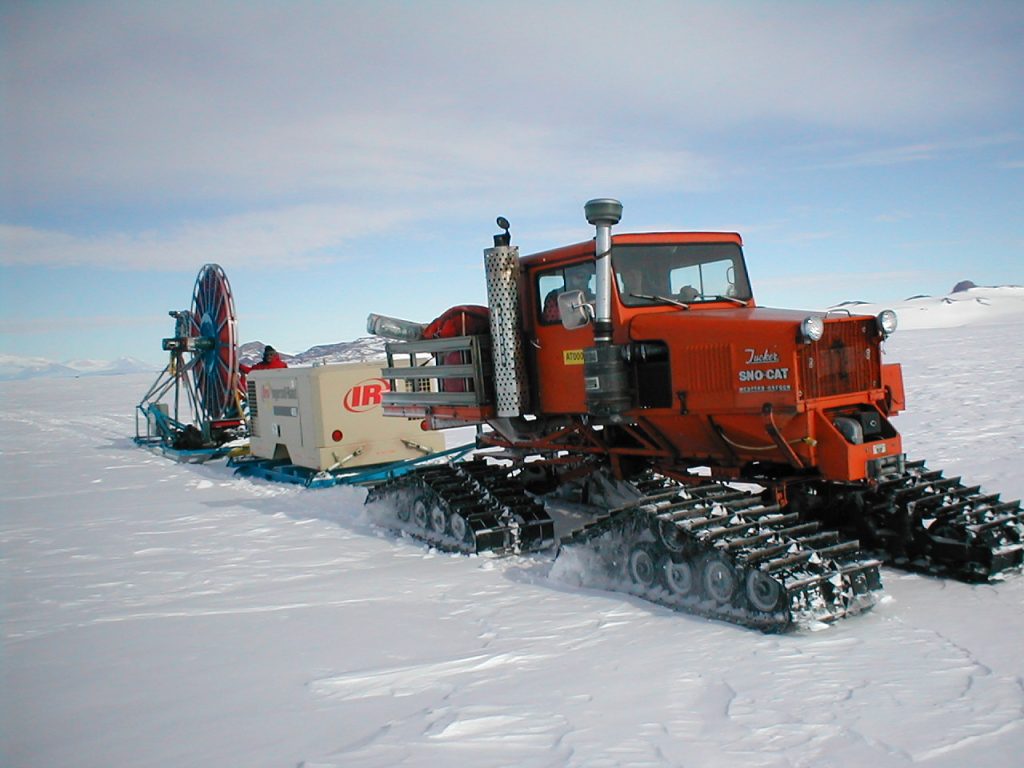 A red snow mobile hauling the shot hole drill on the snow.