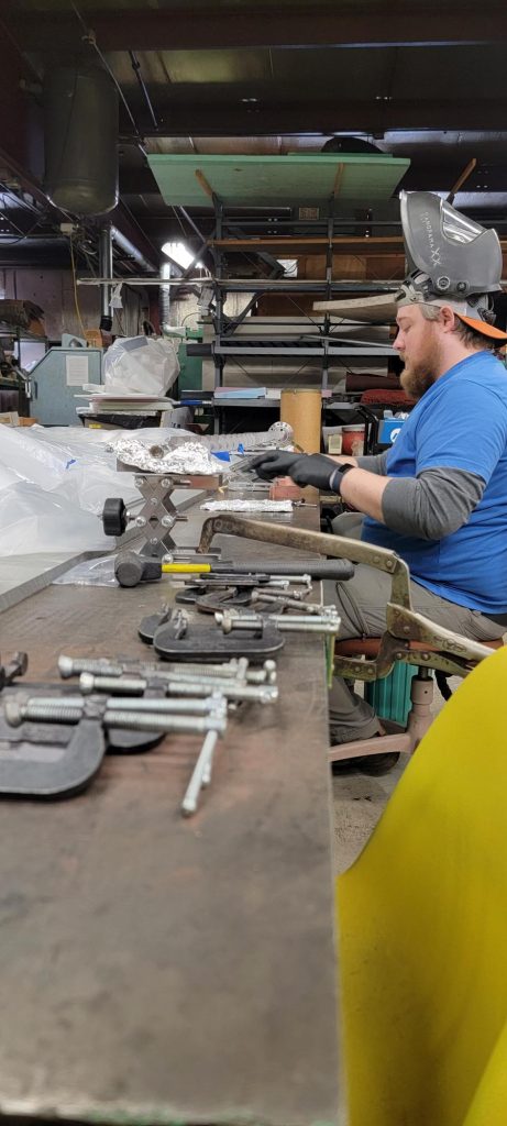 A man with a gray beard and blue shirt wearing a welders mask, working on a metal table with copper blocks.