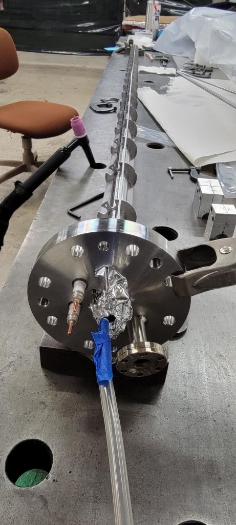 A metal rib for the W7-X project. Closest to the front is a metal disc with holes around the perimeter, with a long metal tube with discs on it.
