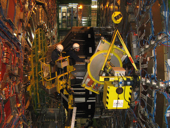 Two people in dark blue jumpsuits working on the inside of the CMS, with a yellow crane.